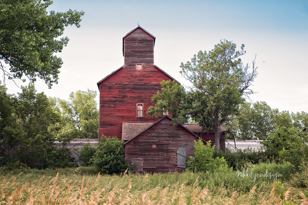 Historic red wooden grain elevator and mill building in rural South Dakota surrounded by tall prairie grass.